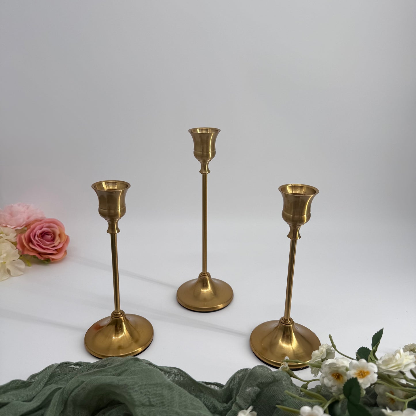 Three brass candlesticks on a white surface with flowers around them