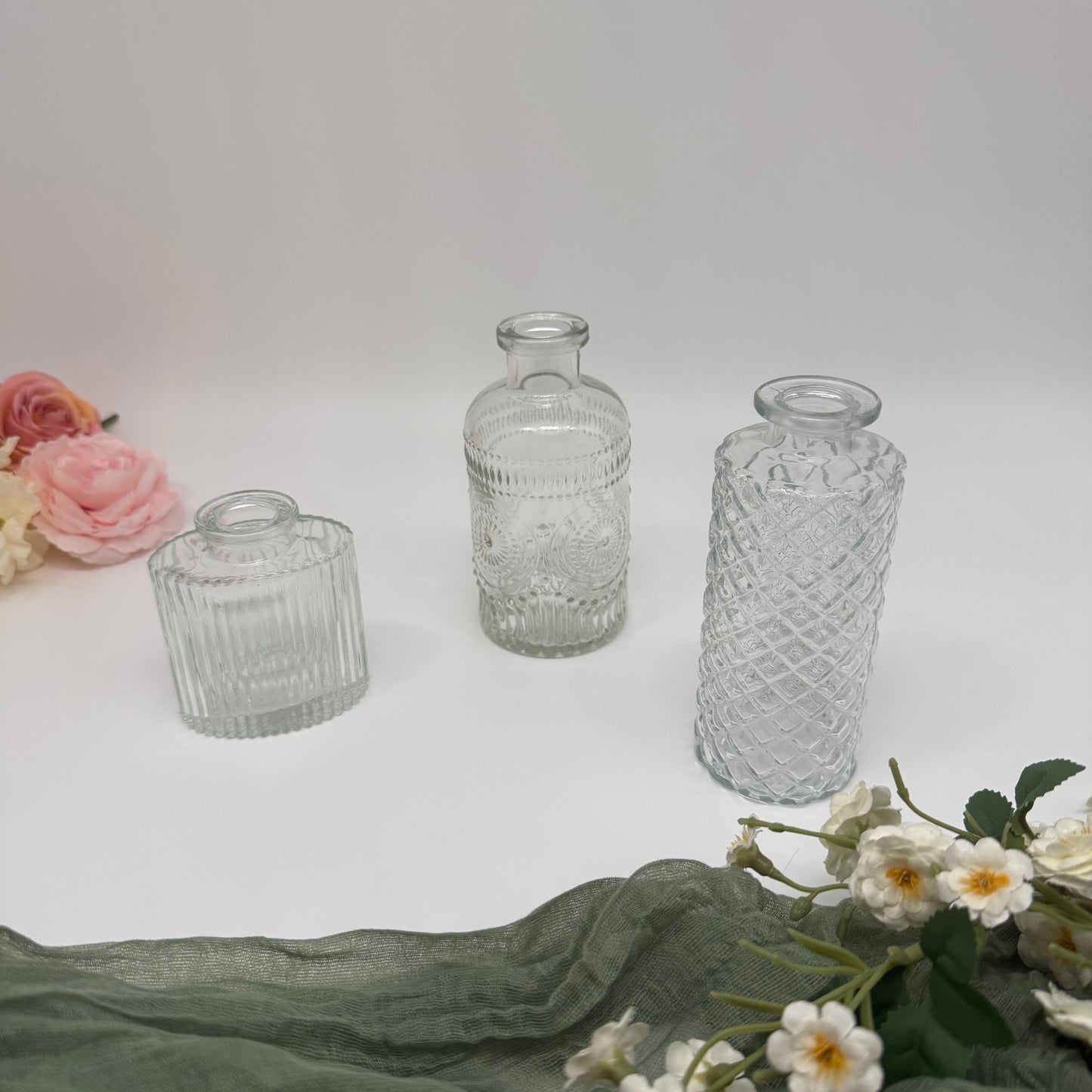 Three clear textured glass bud vases on a light surface with flowers around.