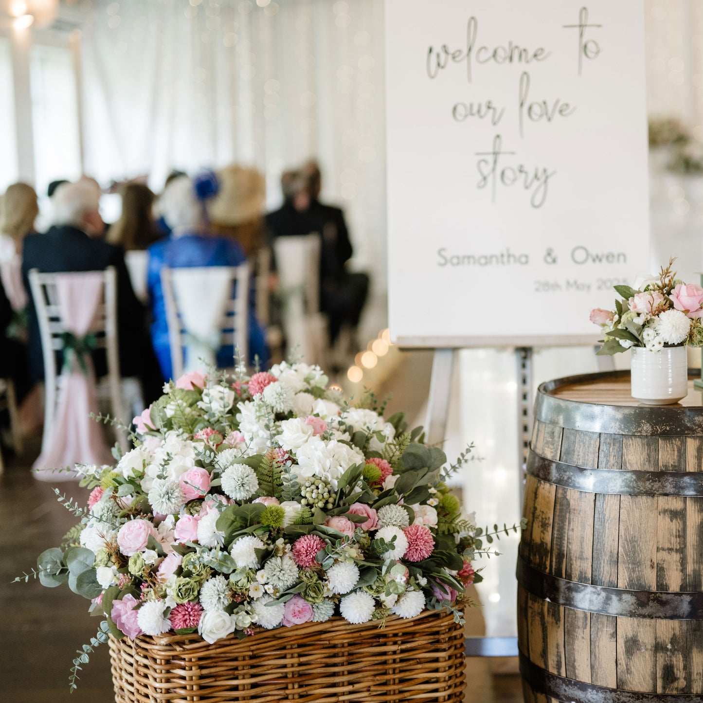 Decorative floral arrangement on a wooden barrel with 'welcome to our love story' sign in the background. Large wicker basket with flower arrangement in foreground.