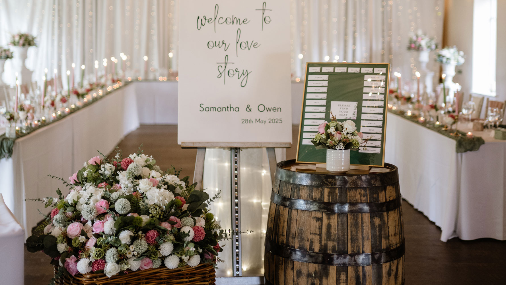Wedding setup with floral arrangements, sign, and barrel table in a decorated hall.
