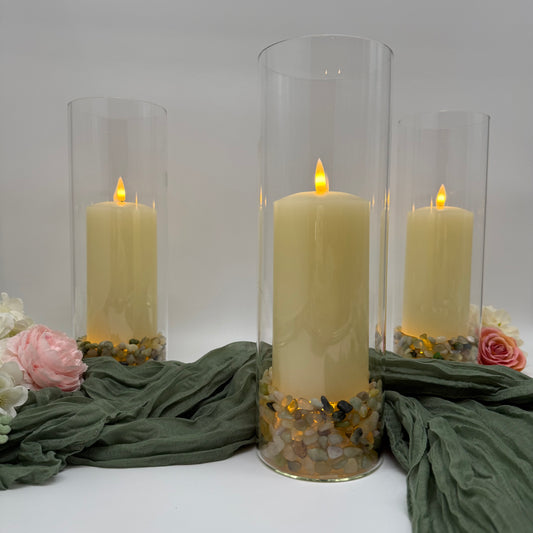 Three lit candles in glass hurricane vase holders with decorative stones and flowers on a white background