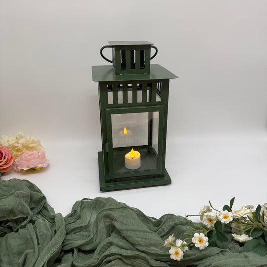 Green lantern with a tea light candle inside on a white background with green fabric and flowers.