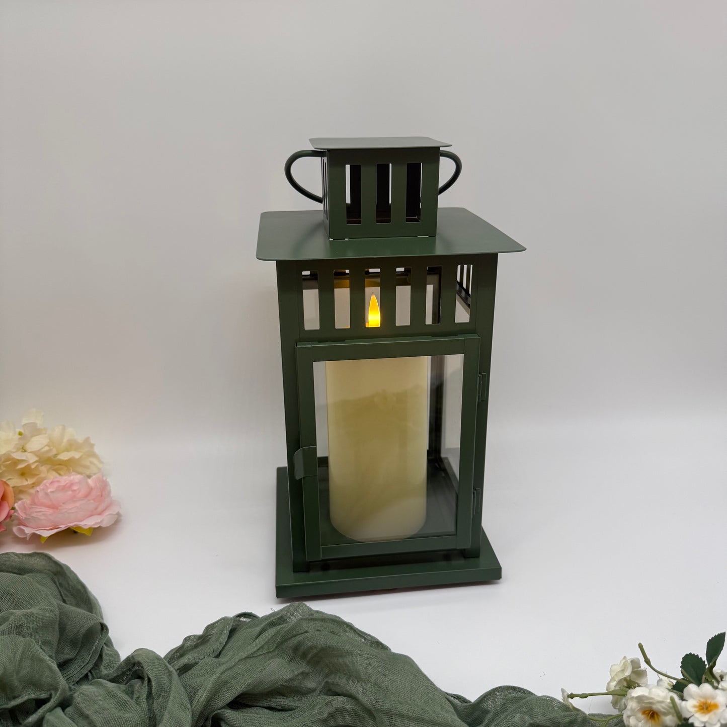Green lantern with a pillar candle on a white background with green fabric and flowers.
