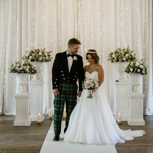 Wedding couple standing in front of a decorative backdrop of flowers in urns and a backdrop of fairy lights and white material.
