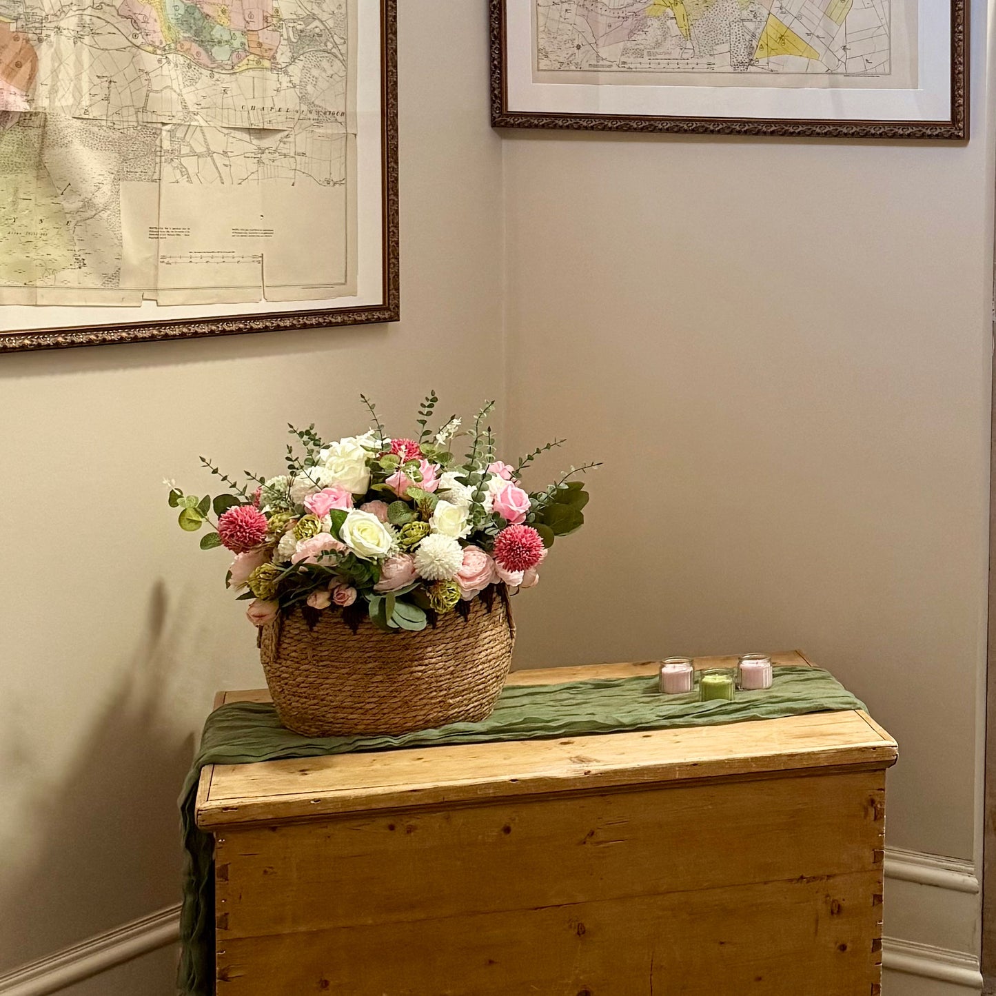 Floral arrangement in a woven basket on a wooden chest with framed maps on the wall.