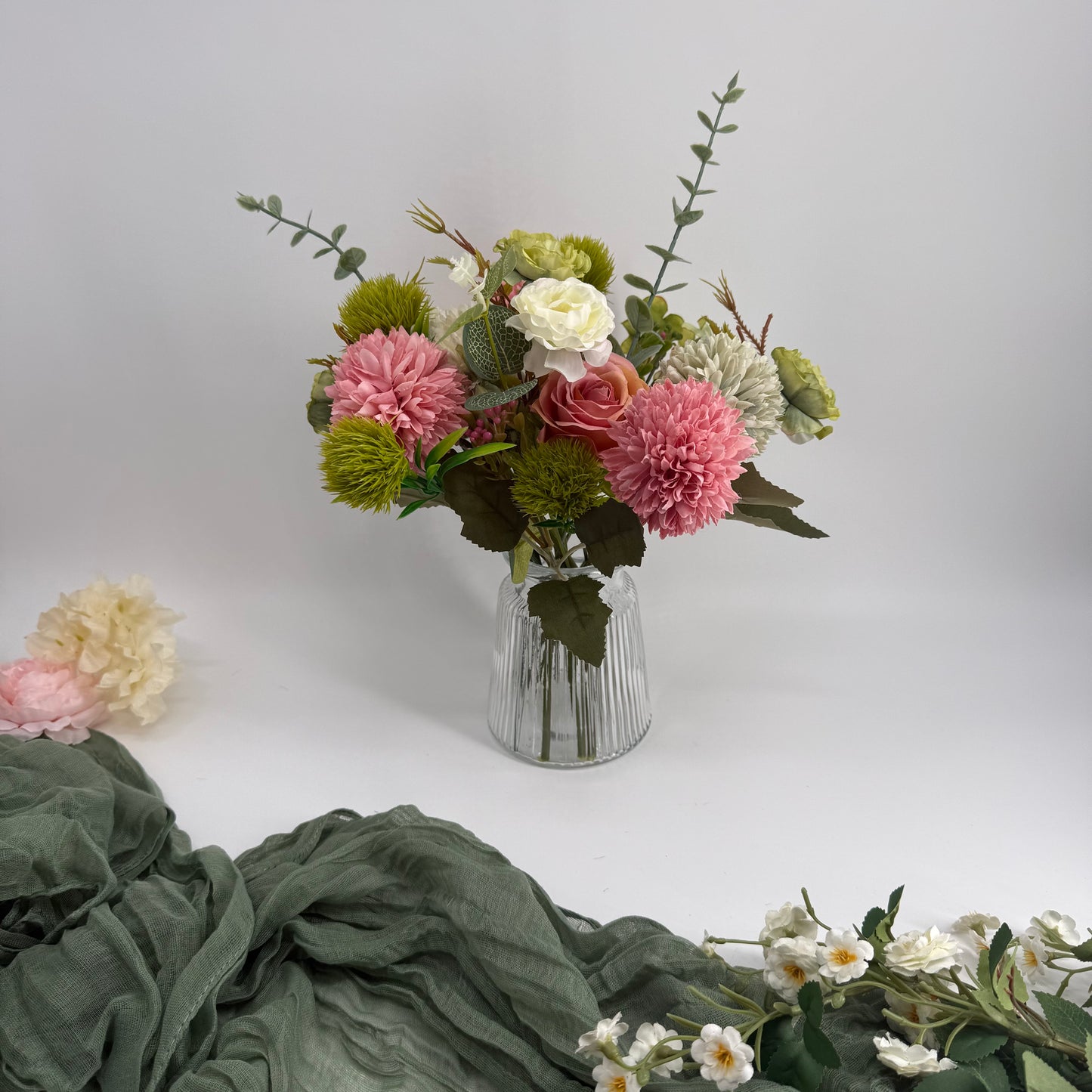 Bouquet of artificial flowers in a clear vase on a white surface with green fabric and additional flowers in the foreground.