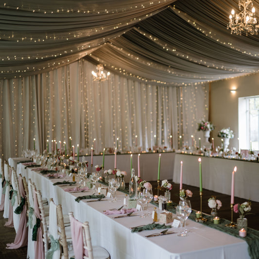 Decorated wedding reception with string lights, candles, and floral arrangements. With seating places set and chair drapes