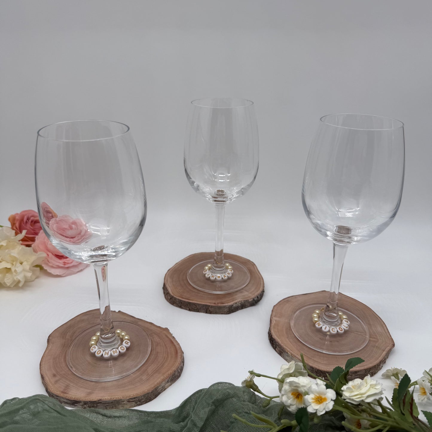 Three wine glasses with wine charms on wooden coasters with flowers around on a white background