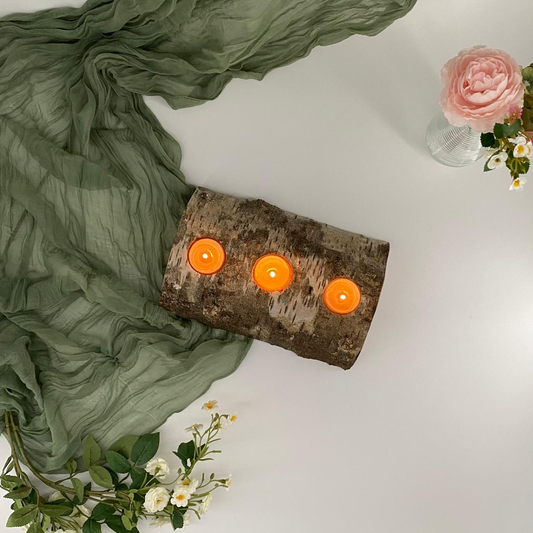 Birch log with three orange candles on a white surface with green fabric and flowers.