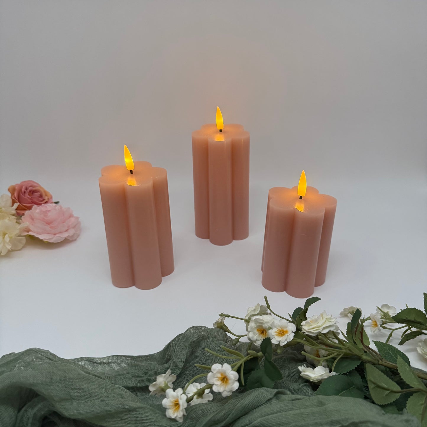 Set of three pink candles with small flames on a white surface with flowers and green fabric.