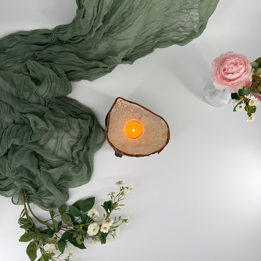 Wooden candle holder with a lit candle, green fabric, and flowers on a white surface