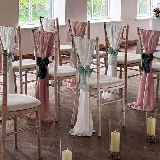 Decorated chairs with pink and white drapes and green ribbons in a room with large windows.