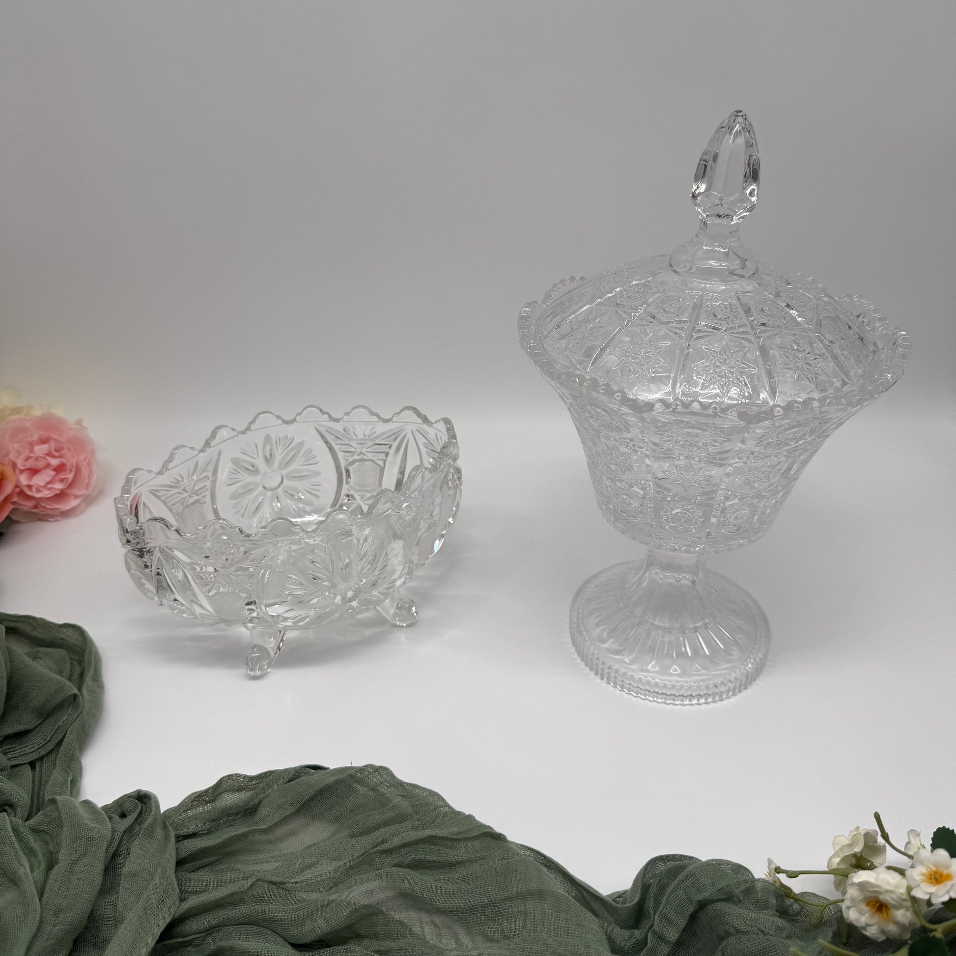 Two clear crystal dishes, a bowl and a covered dish, on a white surface with green fabric and flowers.
