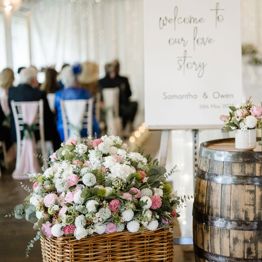 Decorative floral arrangement on a wooden barrel with 'welcome to our love story' sign in the background. Large wicker basket with flower arrangement in foreground.