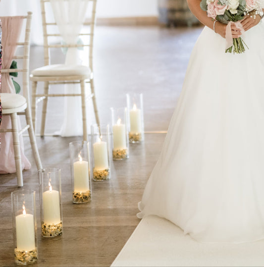 Pillar candles in hurricane vases along the side of a white aisle runner with the bride walking down it.