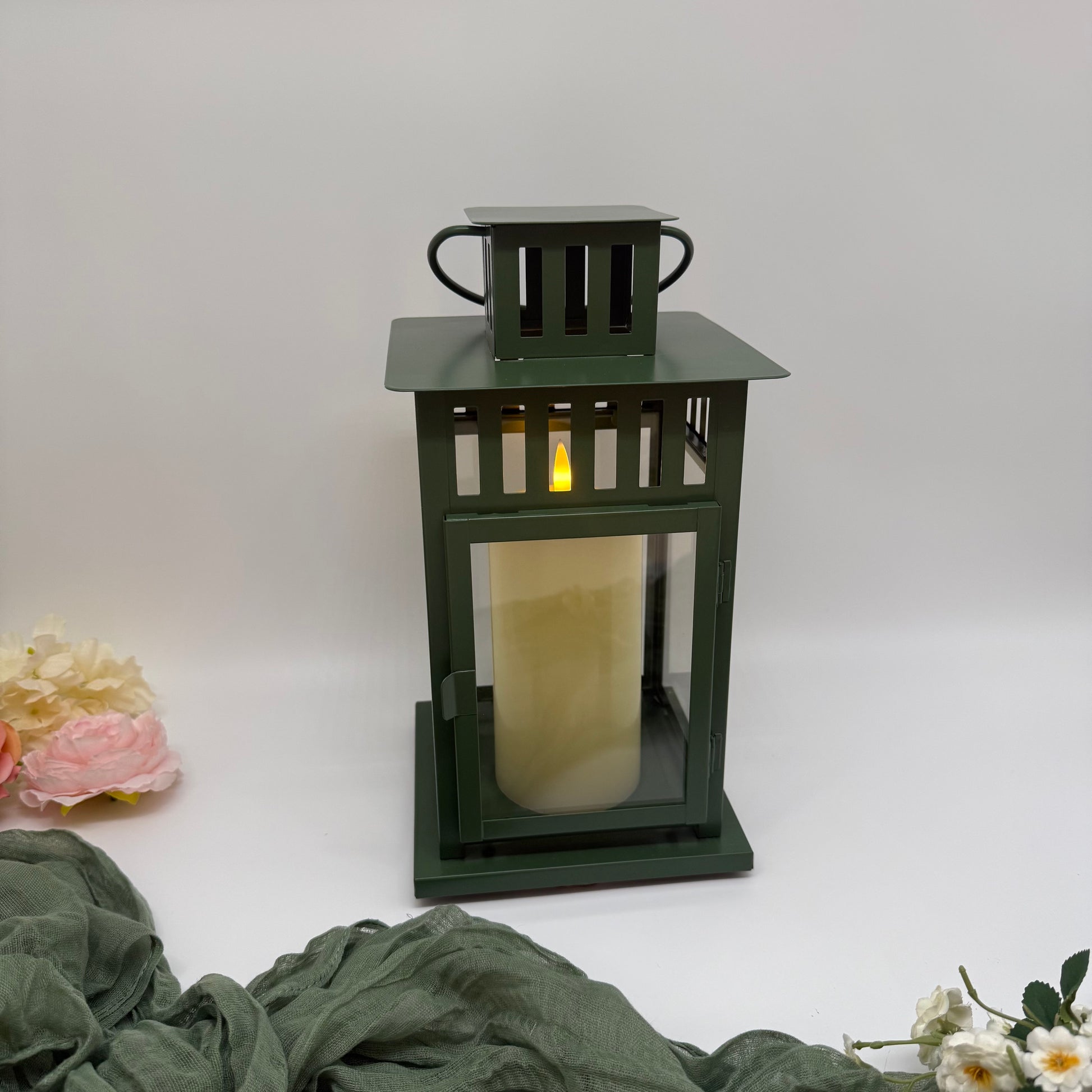 Green lantern with a pillar candle on a white background with green fabric and flowers.