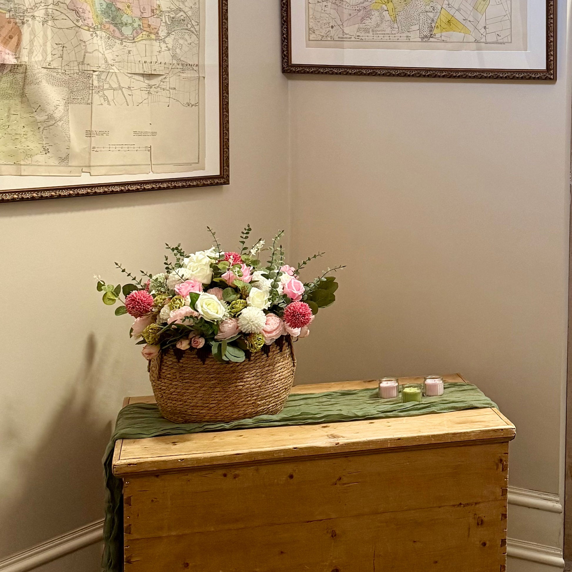 Floral arrangement in a woven basket on a wooden chest with framed maps on the wall.