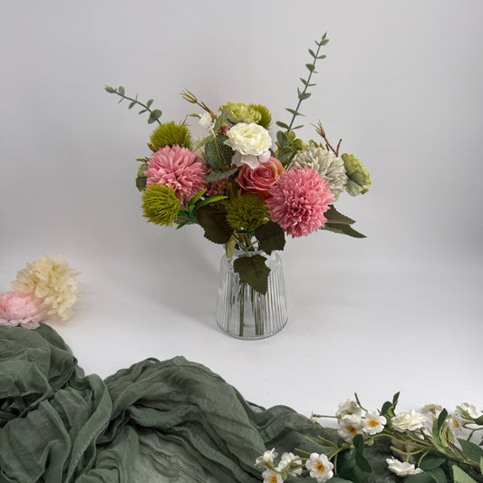 Bouquet of artificial flowers in a clear vase on a white surface with green fabric and additional flowers in the foreground.