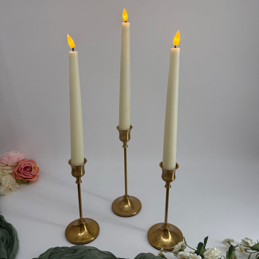 Three lit candles in brass candlesticks on a white surface with flowers.