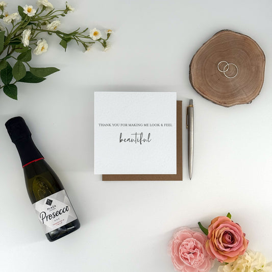 Prosecco bottle, card that says 'thank you for making me look & feel beautiful', pen, and flowers on a white surface