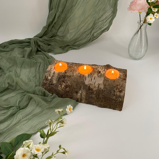 Birch bark candle holder with three orange candles on a white surface with green fabric and flowers.