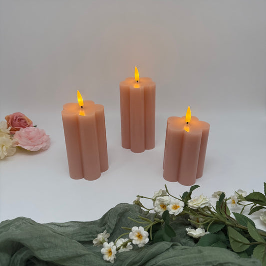 Set of three pink candles with small flames on a white surface with flowers and green fabric.