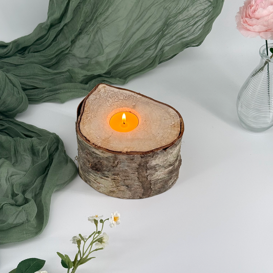 Wooden candle holder with a lit candle on a white surface with green fabric and flowers.